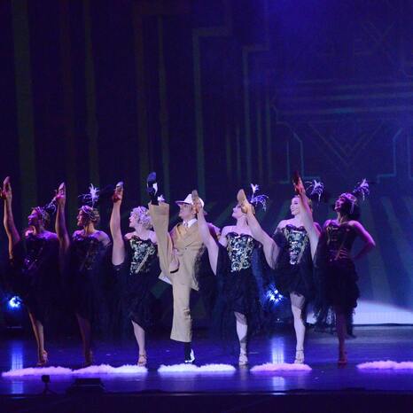 Es sind sieben Tänzer und Tänzerinnen auf der Bühne. Sie tragen 20er Jahre Looks. Sie stehen in einer Reihe zum Publikum. Halten sich aneinander fest und werfen jeweils das linke Bein hoch. So dass sie quasi einen Spagat im stehen vollführen.