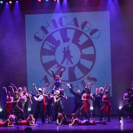 Das Odyssey Dance Theater steht auf der Bühne. Es sind 21 Menschen zu sehen. Alle tragen chice Outfits im zwanziger Jahre Look. Die Tanztruppe steht gerade in einem Standbild. Im Hintergrund hängt ein Banner mit der Aufschrift "Chicago Nights".