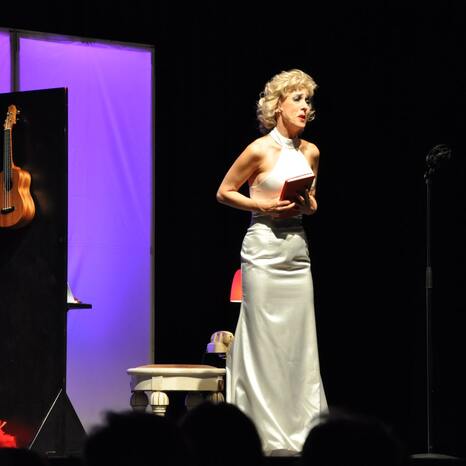 Eine Schauspielerin steht auf der Bühne. Sie soll Marilyn Monroe verkörpern mit blonden Locken, Make up und einem weißen, langen, schicken Kleid. Sie hält ein Buch in der Hand und singt zum Publikum. Auf der Bühne ist es sehr dunkel, so dass das weiße Kleid strahlt.