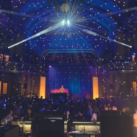 Der Theatersaal wird durch eine Discokugel hell erleuchtet. Menschen tanzen zur Musik. Ein DJ legt auf.